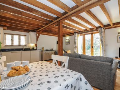An open plan living area with wooden ceiling beams a kitchen with cupboards a dining table with croissants and mugs and a sitting area with gray sofas and glass doors at Stow Cottage in Davidstow near Camelford
