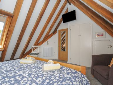 A bedroom with wooden beams on the ceiling a bed with floral bedding a white chair a wall-mounted television and a brown armchair at Stow Cottage in Davidstow near Camelford