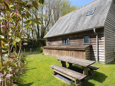 An outdoor picnic table and benches on grass next to a wooden building with trees in the background at Stow Cottage in Davidstow near Camelford