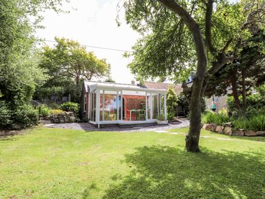 A garden with a lawn tree and a glass conservatory with colorful chairs at Morawel in Newport Pembrokeshire