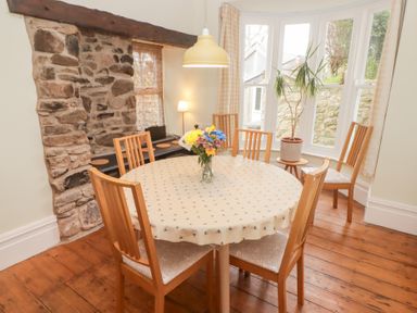 A dining room with a round table covered with a tablecloth and wooden chairs near a stone wall and windows at Morawel in Newport Pembrokeshire