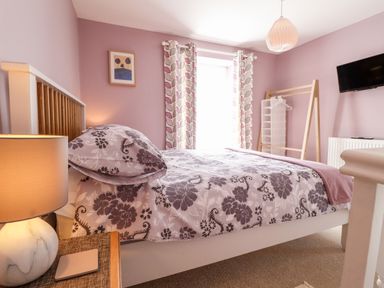 A bedroom with a bed with floral bedding a lamp on a bedside table patterned curtains a wall-mounted TV and a wooden clothes rack at Morawel Fach in Newport Pembrokeshire