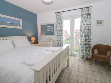 A bedroom with a white bed a dresser with a lamp and an alarm clock a wicker chair and windows with patterned curtains at Silversands in Filey