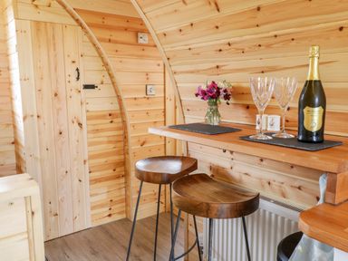 A wooden interior with two bar stools a counter with a bottle of prosecco two glasses and a small vase with flowers at Embden Pod at Banwy Glamping in Llanfair Caereinion