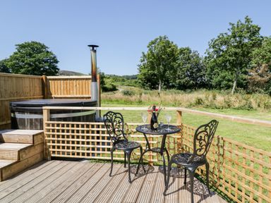 An outdoor wooden deck with a small black metal table and two chairs a hot tub with steps and wooden fencing with trees and grass in the background at Embden Pod at Banwy Glamping in Llanfair Caereinion