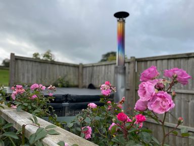 Outdoor area with pink flowers and a hot tub with a multicolored chimney behind a wooden fence at Embden Pod at Banwy Glamping in Llanfair Caereinion