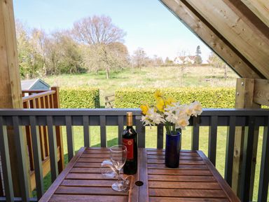 A balcony with a wooden table holding a wine bottle two wine glasses and a vase with flowers overlooking a garden at Cherrywood Barn in South Petherton