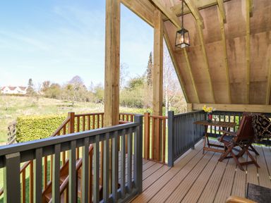 A wooden covered balcony with railing a small table and two chairs overlooking a grassy area and trees at Cherrywood Barn in South Petherton