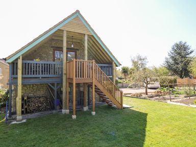 A stone house with a wooden balcony and stairs overlooking a garden and driveway at Cherrywood Barn in South Petherton