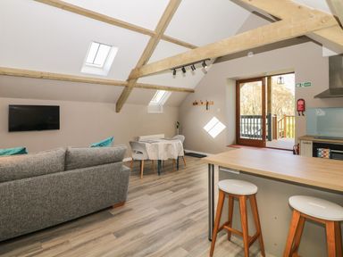 An open living space with a gray sofa a small dining table two white chairs kitchen counter with two stools and wooden beams on the ceiling at Cherrywood Barn in South Petherton