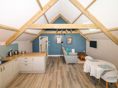 An open plan room with a kitchen area, sofa, coffee table, dining table and chairs under exposed wooden beams at Cherrywood Barn in South Petherton