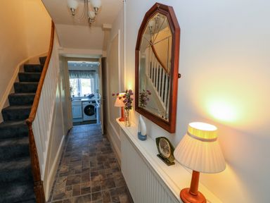 A hallway with a staircase and a shelf with a lamp clock mirror and vase leading to a kitchen area at Ty Coch y Castell in Pembroke