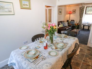 A dining table set with plates glasses cutlery and a vase with flowers next to a living room with leather sofas and a lamp at Ty Coch y Castell in Pembroke
