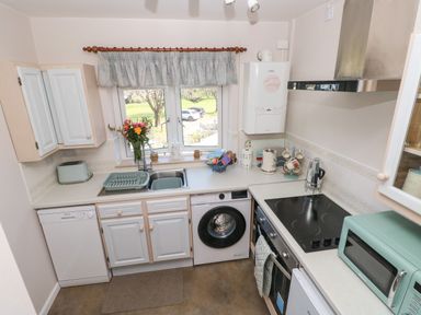A kitchen with white cabinets a washing machine a dishwasher a toaster a microwave and a window with a curtain at Ty Coch y Castell in Pembroke