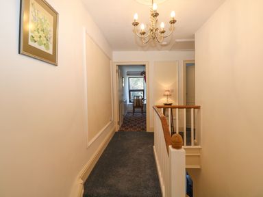 A hallway with carpeted floor a chandelier a framed picture and a wooden stair railing at Ty Coch y Castell in Pembroke