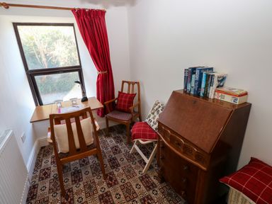 A small room with a wooden table and two chairs near a window with red curtains and a wooden cabinet with books on top at Ty Coch y Castell in Pembroke