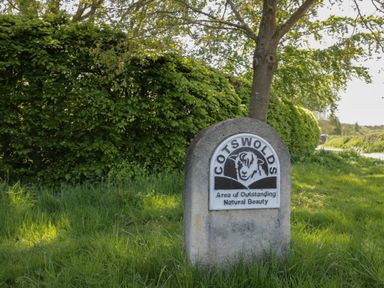 A stone sign with a sheep illustration and text about Cotswolds Area of Outstanding Natural Beauty by a tree and hedge