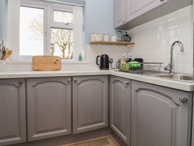 A kitchen corner with grey cabinets wooden cutting boards on a windowsill a black kettle and a dish rack at Molly's Bit Church Westcote near Bourton-On-The-Water