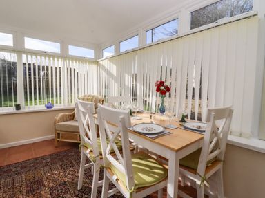A dining area with a wooden table set for four and white chairs with green cushions in a sunroom with vertical blinds at Molly's Bit Church Westcote near Bourton-On-The-Water
