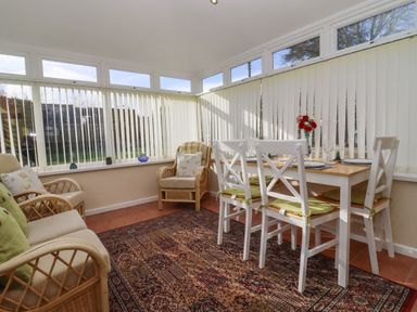 A sunroom with wicker chairs a dining table with white chairs and a patterned rug at Molly's Bit in Church Westcote near Bourton-On-The-Water