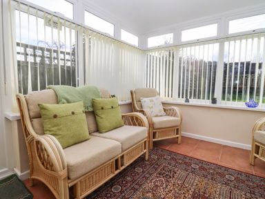 A sunroom with wicker furniture including a two-seater and armchairs with green and floral cushions and a patterned rug on tiled floor at Molly's Bit in Church Westcote near Bourton-On-The-Water