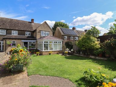 A garden with grass, flowers, and a seating area behind a house with a conservatory at Mollys Bit in Church Westcote near Bourton-On-The-Water