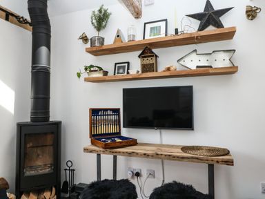 A room with a black wood stove a wall mounted TV two wooden shelves with decorations and a wooden table with silverware case and placemat at Rocket Cart House in Lizard