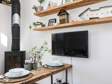 A dining area with wooden table set for two under wall mounted shelves and a flat screen TV at Rocket Cart House in Lizard
