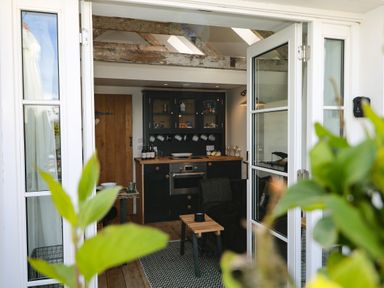 View through open glass doors into a small kitchen with black cabinets wooden countertop and wooden door at Rocket Cart House in Lizard