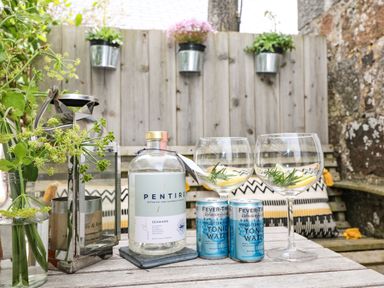 A wooden table with a bottle of Pentire Seaward, two cans of Fever-Tree tonic water, two glasses with lemon and herbs, a lantern, and flowers with a wooden fence and bench in the background at Rocket Cart House in Lizard
