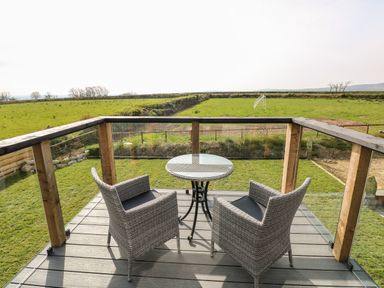A balcony with two wicker chairs and a round glass table overlooking a grassy field at Glowty Brynbedw in Efailwen