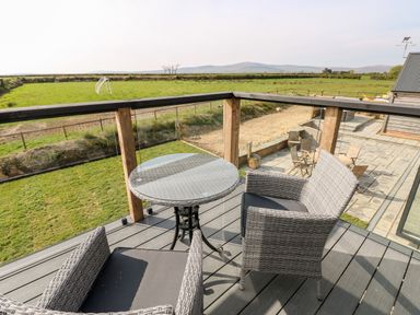 An outdoor balcony with a round glass table and two wicker chairs overlooking a grassy field and a patio with wooden chairs at Glowty Brynbedw in Efailwen