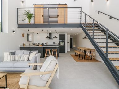 An open plan living area with a gray sofa and chair a kitchen with island and bar stools a dining table with benches and a staircase with metal railing at Tilia Barn in Wincanton