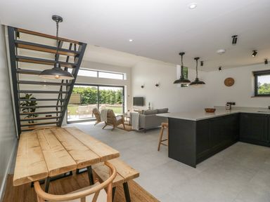 An open plan living area with a wooden dining table and bench under a staircase a sofa and armchairs facing a wall mounted TV and a kitchen island with bar stools at Tilia Barn Wincanton