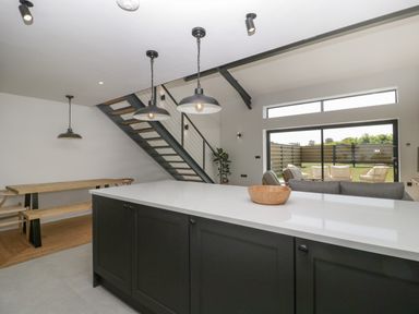 A kitchen island with a wooden bowl and three pendant lights above a dining area with a wooden table and benches near a staircase and a living area with chairs seen through large sliding glass doors at Tilia Barn in Wincanton