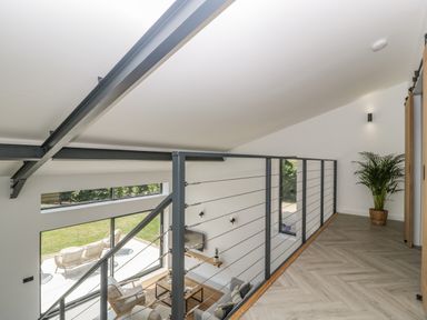 A modern interior view showing a mezzanine with metal railing and a plant in a pot overlooking a living area with seating at Tilia Barn in Wincanton