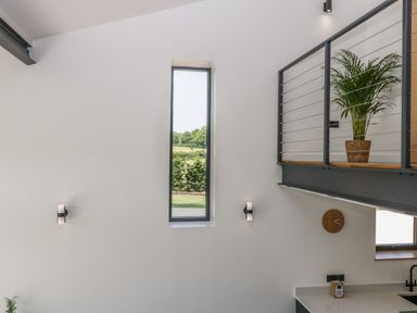 An interior space with a tall narrow window two wall lights a clock a plant on an upper railing and part of a kitchen counter at Tilia Barn in Wincanton