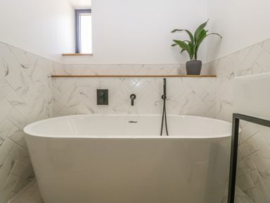 A bathroom with a freestanding bathtub a small window a plant on a wooden shelf and marble tiled walls at Tilia Barn in Wincanton