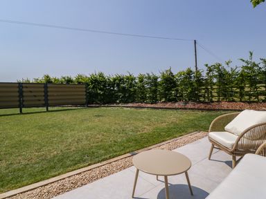 An outdoor patio area with a round table and cushioned chair next to a lawn and hedge at Tilia Barn in Wincanton