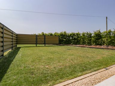 A small grassy backyard with a beige and black fence and a row of green bushes at Tilia Barn in Wincanton