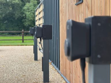 A row of black charging stations on metal stands next to a wooden building exterior with a green grassy area and wooden fence in the background at Tilia Barn in Wincanton