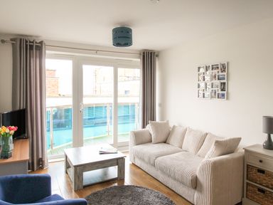 A living room with a white sofa, wooden coffee table, blue armchair, TV on a stand, and glass doors leading to a balcony at Nautical Breeze in Weymouth