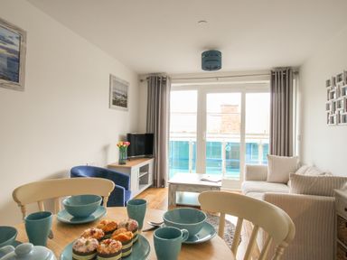 A dining table with blue dishes and muffins near a living room with sofa, armchair, coffee table, TV, and large windows at Nautical Breeze in Weymouth
