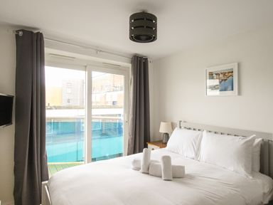 A bedroom with a bed with white linens and rolled towels a bedside table with a lamp a wall-mounted television and sliding glass doors with curtains at Nautical Breeze Weymouth