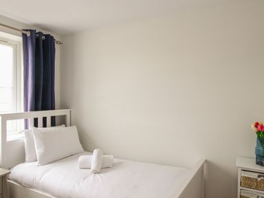 A single bed with white bedding next to a window with blue curtains and a small bedside table with flowers at Nautical Breeze in Weymouth