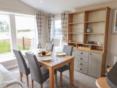 A dining area with a wooden table and four grey chairs beside windows and a wooden cabinet at Sea Holly Lodge in Runswick Bay