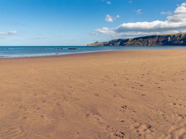 A sandy beach with footprints and a calm sea with cliffs in the distance at Sea Holly Lodge in Runswick Bay