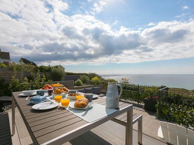 An outdoor dining table with breakfast items and drinks on a patio overlooking the sea at Sea Sands in Praa Sands