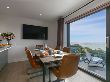 A dining area with a table set for breakfast and chairs next to a large window overlooking the sea at Sea Sands in Praa Sands