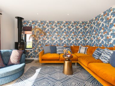A living room with an orange corner sofa blue armchair patterned cushions a black fireplace and a blue and white rug at Sea Sands in Praa Sands
